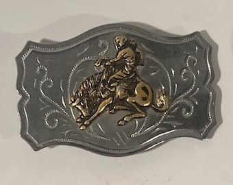 May include: Silver belt buckle with a gold-toned design of a cowboy riding a bucking bronco. The buckle has a detailed floral pattern around the edges.