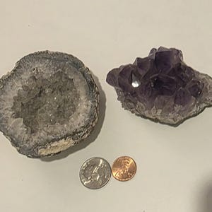 May include: An open geode with crystal formations and a cluster of amethyst crystals. A quarter and a penny are next to the crystals for size comparison. The geode is gray and white, and the amethyst is a deep purple.