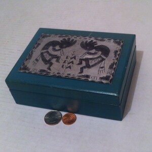 May include: A teal blue wooden box with a silver inlay depicting two Native American dancers. The box has a hinged lid and is closed.