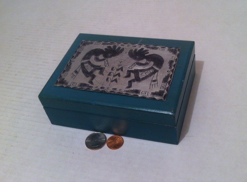 May include: A teal blue wooden box with a silver inlay depicting two Native American dancers. The box has a hinged lid and is closed.