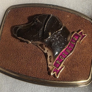 May include: A rectangular belt buckle with a brown suede-like background and a gold-colored border. The buckle features a raised image of a dog's head in black and white, with a pink banner that reads "POINTER".