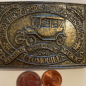May include: A gold-toned metal belt buckle featuring a vintage Ford Model T car with the text "Heavy Ford Improvement Record Model T Ford Automobiles Year 25".