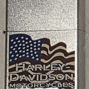 May include: A silver-colored lighter with a textured surface. The lighter features a design of the American flag with the words "HARLEY-DAVIDSON MOTORCYCLES" in a brown font.