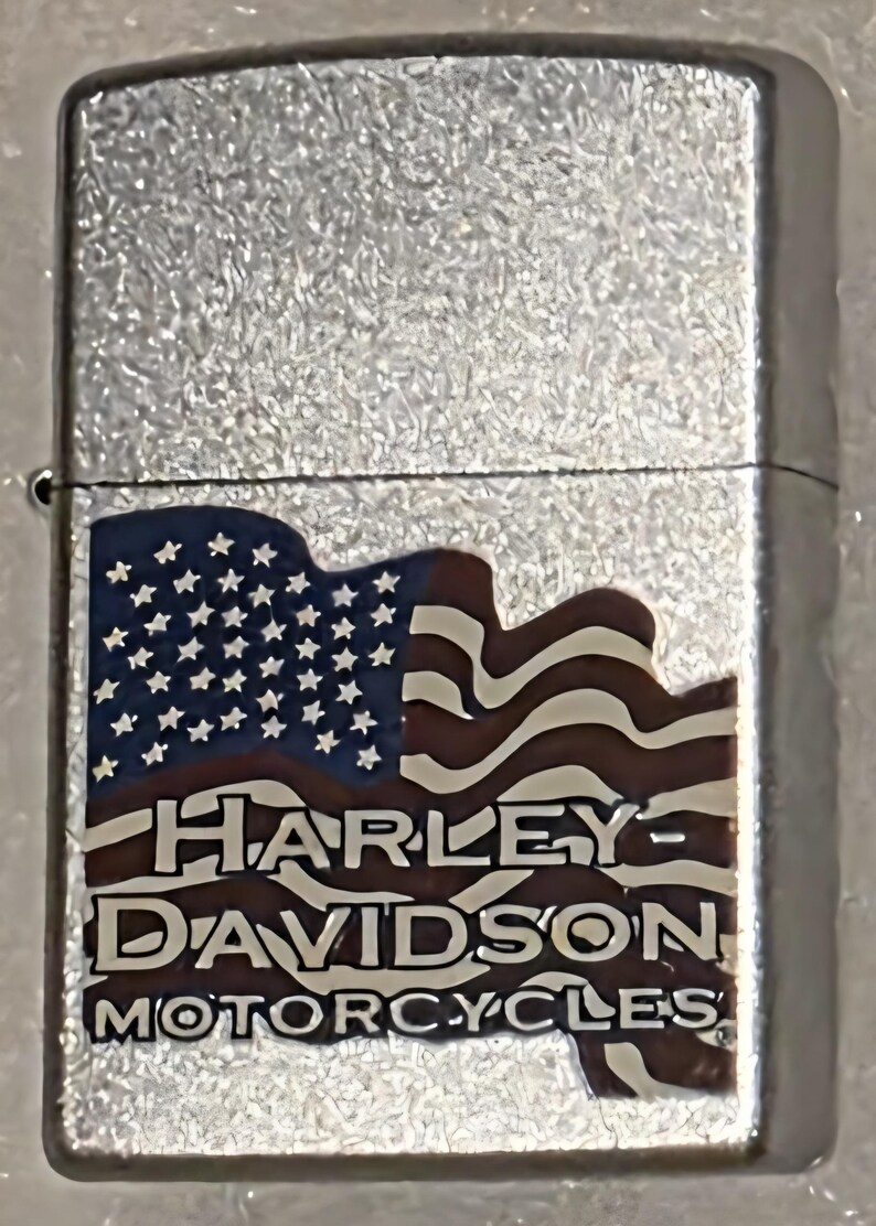 May include: A silver-colored lighter with a textured surface. The lighter features a design of the American flag with the words "HARLEY-DAVIDSON MOTORCYCLES" in a brown font.