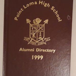 May include: A maroon leather-bound book with gold lettering that reads "Point Loma High School" and "Alumni Directory 1999". The book features a gold crest with a shield and a compass.