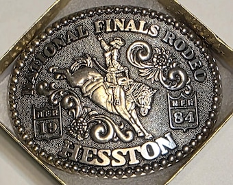 May include: An oval silver-toned belt buckle featuring a raised relief of a person riding a bucking horse. The text reads NATIONAL FINALS RODEO, HESSTON, and NFR 1984. The border is decorated with a beaded pattern and scrollwork accents.