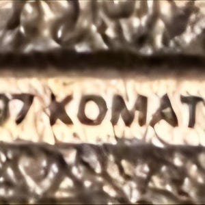 Op de afbeelding: Close-up van een getextureerd, beige oppervlak met een rechthoekige inzet. De inzet toont het copyrightsymbool, het jaar "1997" en de naam "KOMATSU" in bruine letters. De algehele uitstraling suggereert een vintage of verouderd item.