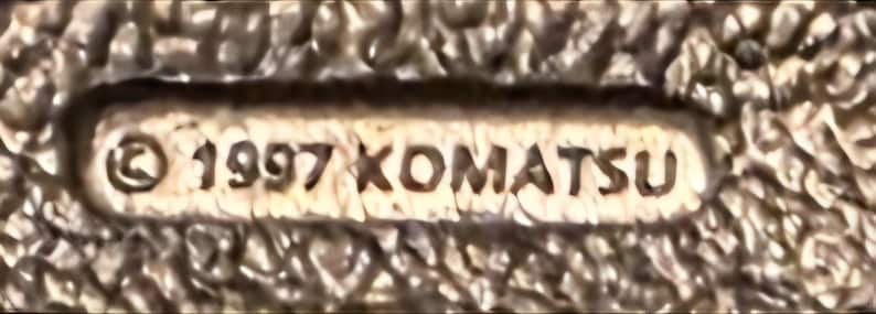 Op de afbeelding: Close-up van een getextureerd, beige oppervlak met een rechthoekige inzet. De inzet toont het copyrightsymbool, het jaar "1997" en de naam "KOMATSU" in bruine letters. De algehele uitstraling suggereert een vintage of verouderd item.