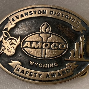 Puede incluir: Una hebilla de cinturón de metal de color dorado con fondo negro. La hebilla presenta el texto "Evanston District", "Amoco", "Wyoming" y "Safety Award". La hebilla también presenta una representación estilizada de una plataforma petrolera, una llama y un animal estilizado.