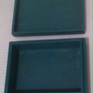 May include: A teal blue rectangular box with a hinged lid. The box is empty and is sitting on a white surface. Two coins are in front of the box.