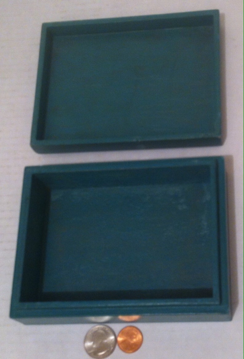 May include: A teal blue rectangular box with a hinged lid. The box is empty and is sitting on a white surface. Two coins are in front of the box.