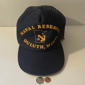 May include: A navy blue trucker hat with a yellow embroidered design. The design features the text "NAVAL RESERVE DULUTH, MINN." and an anchor inside the outline of the state of Minnesota.