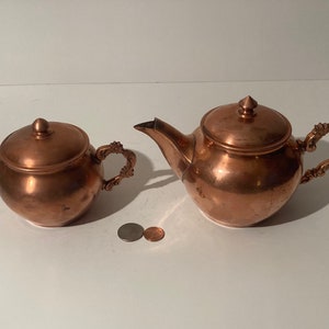 May include: A copper tea set, including a teapot and a sugar bowl, with ornate handles and lids. The teapot has a spout and a rounded body. The sugar bowl is smaller and round. Two coins are next to the tea set.