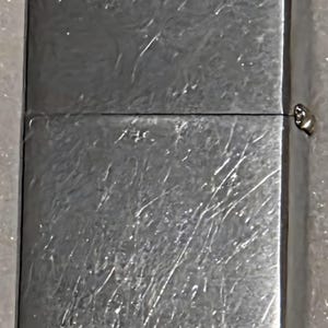 May include: A silver-colored, rectangular lighter with a brushed metal finish. The lighter has a hinged lid and a small, metallic hinge. The surface shows visible scratches and wear, indicating use.