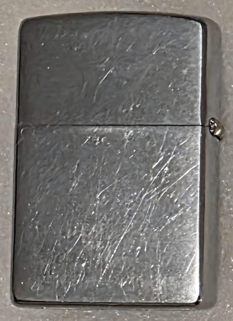 May include: A silver-colored, rectangular lighter with a brushed metal finish. The lighter has a hinged lid and a small, metallic hinge. The surface shows visible scratches and wear, indicating use.