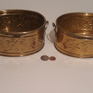 Może przedstawiać: Dwa złote metalowe doniczki z uchwytami. Doniczki mają ozdobny tłoczony wzór winogron i liści.