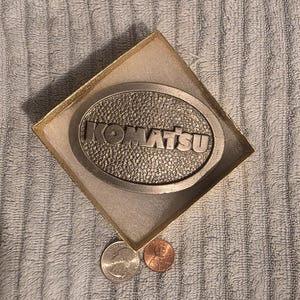 Op de afbeelding: Een zilverkleurige riemgesp met het woord "KOMATSU" in verhoogde letters, geplaatst in een ovale vorm. De gesp zit in een goudkleurige doos. Een kwartje en een penny liggen naast de doos.