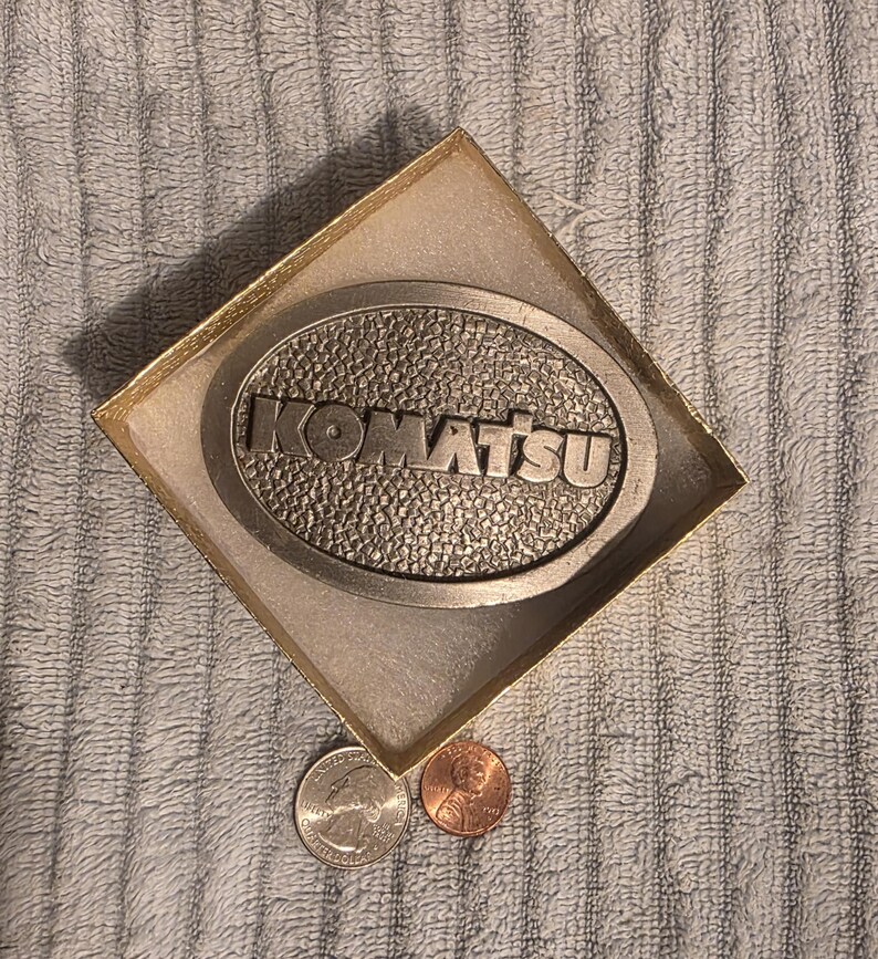 Op de afbeelding: Een zilverkleurige riemgesp met het woord "KOMATSU" in verhoogde letters, geplaatst in een ovale vorm. De gesp zit in een goudkleurige doos. Een kwartje en een penny liggen naast de doos.