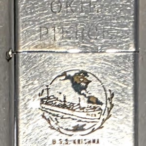 May include: Silver-toned metal lighter with etched design. The lighter features the text "OKIE PIERCE" at the top and a circular emblem with a ship and globe. The text "U.S.S. KOZUMI ARC 3" is at the bottom.