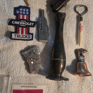 May include: A collection of vintage items including a Chevrolet Trucks patch, a Coca-Cola patch, a silver vase, a bottle opener, a bear-topped bell, a crystal, a corkscrew, a toy cannon, a brass tool, and coins.