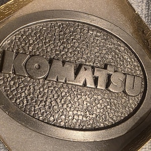Op de afbeelding: Ovale metalen riemgesp met het woord "KOMATSU" in verhoogde letters. De gesp heeft een getextureerde achtergrond en een zilveren afwerking. De gesp zit in een goudkleurige doos.