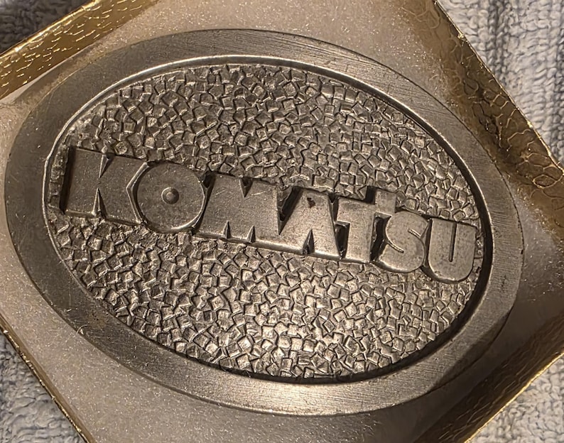 Op de afbeelding: Ovale metalen riemgesp met het woord "KOMATSU" in verhoogde letters. De gesp heeft een getextureerde achtergrond en een zilveren afwerking. De gesp zit in een goudkleurige doos.
