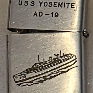 以下が含まれることがあります： 上部に「USS YOSEMITE AD-19」の文字が刻まれた銀色のライター。下半分には船の彫刻が施されています。ライターは開いており、内部が見えます。