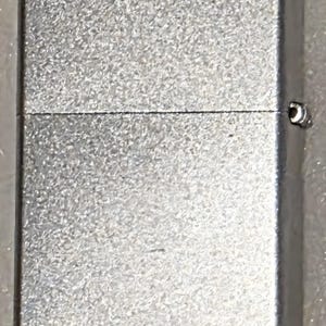 May include: A silver-colored lighter with a textured surface. The rectangular lighter has a hinged lid and a small metal loop on the side. The lighter is a classic design.