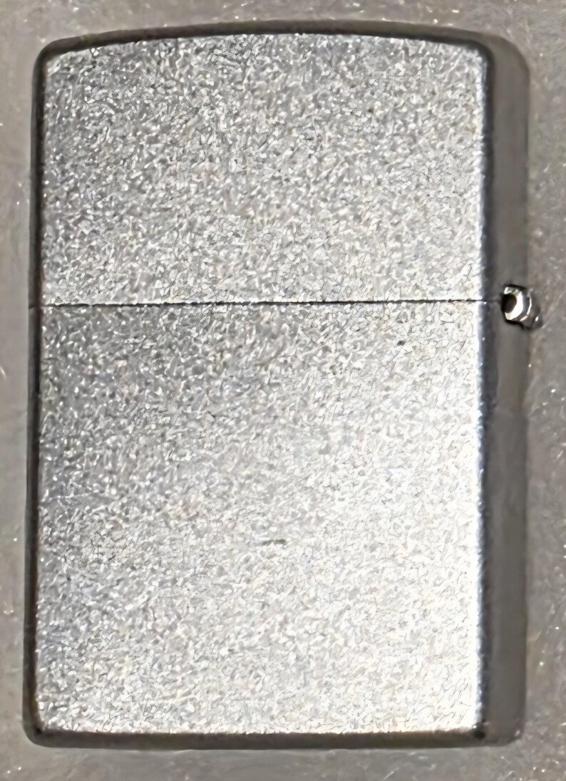 May include: A silver-colored lighter with a textured surface. The rectangular lighter has a hinged lid and a small metal loop on the side. The lighter is a classic design.