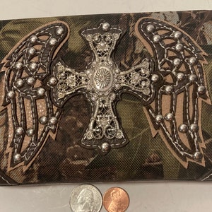 May include: A brown and green camouflage fabric wallet with a silver cross and wings with rhinestones. The wallet has a brown leather trim.