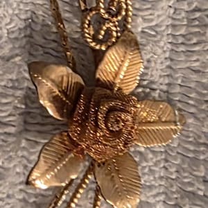 Puede incluir: Un collar dorado con un colgante floral. El colgante presenta un diseño de flor con hojas detalladas y una espiral central. La cadena del collar es delicada y complementa el diseño floral. La joya está sobre un fondo texturizado.