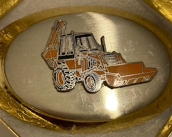 Puede incluir: Hebilla de cinturón ovalada con un diseño detallado de una retroexcavadora Case 580 naranja y negra. La hebilla tiene un centro plateado y un borde dorado texturizado, presentada en una caja dorada.