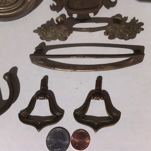 May include: Assortment of antique brass hardware, including two ornate door pulls, a decorative handle, and a long, curved pull. The hardware is arranged on a white surface with two US coins for scale.