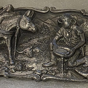 May include: Silver-toned metal belt buckle featuring a detailed relief scene of a prospector panning for gold. A donkey with saddle stands near a mountain landscape. The buckle has an ornate, decorative border.