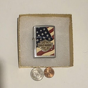 May include: A silver Harley-Davidson lighter with an American flag design, including red and white stripes and white stars on a blue background. The lighter is in a small cardboard box with two coins.