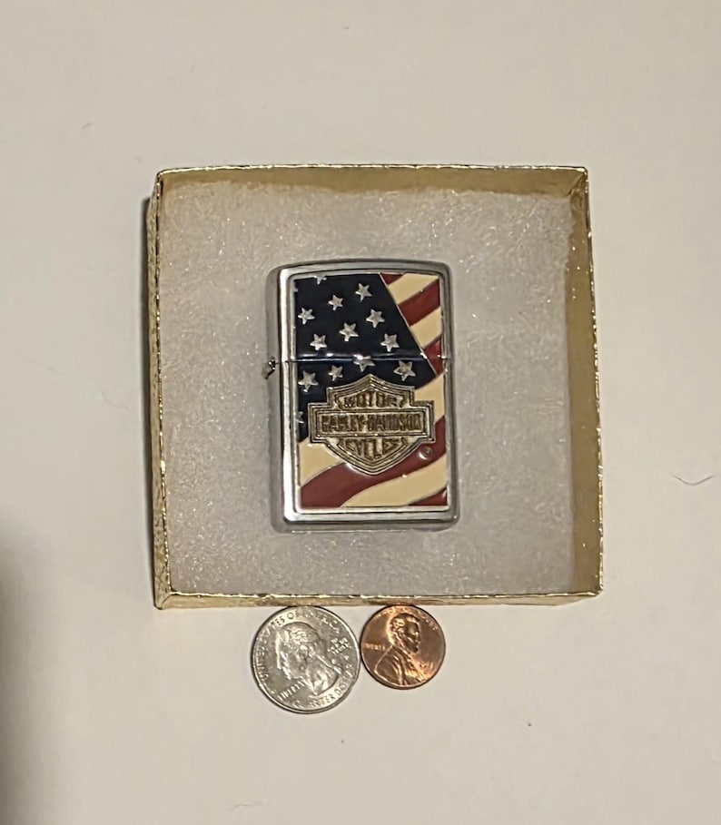 May include: A silver Harley-Davidson lighter with an American flag design, including red and white stripes and white stars on a blue background. The lighter is in a small cardboard box with two coins.