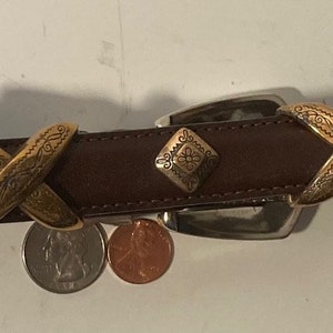 Puede incluir: Un cintur&oacute;n de cuero marr&oacute;n con detalles met&aacute;licos dorados. El cintur&oacute;n tiene un patr&oacute;n decorativo en X en los detalles met&aacute;licos y una hebilla plateada.