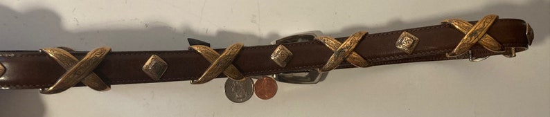 Puede incluir: Un cintur&oacute;n de cuero marr&oacute;n con detalles met&aacute;licos dorados. El cintur&oacute;n tiene un patr&oacute;n decorativo en X en los detalles met&aacute;licos y una hebilla plateada.