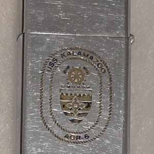 May include: Silver-toned metal lighter with a brushed finish. The lighter features an engraved emblem with the text "USS KALAUA CO" and "AOR-6". The emblem includes a ship's wheel and a shield design.