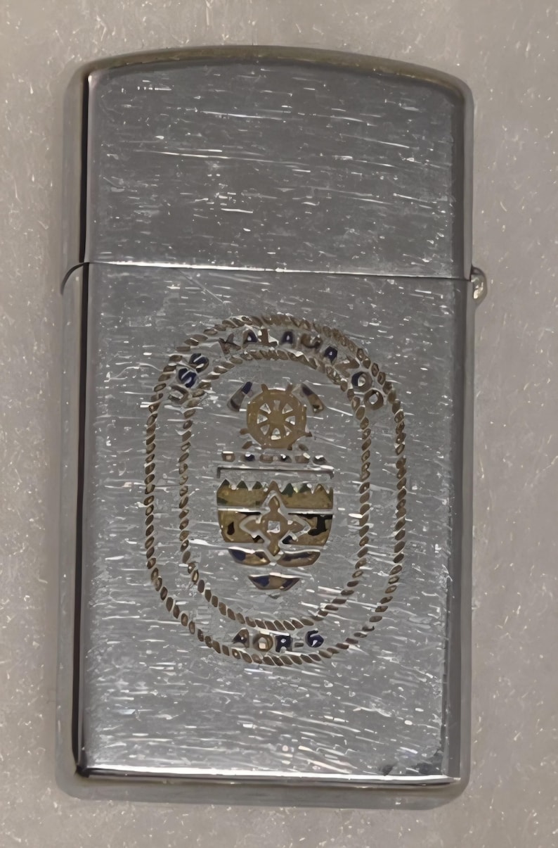 May include: Silver-toned metal lighter with a brushed finish. The lighter features an engraved emblem with the text "USS KALAUA CO" and "AOR-6". The emblem includes a ship's wheel and a shield design.