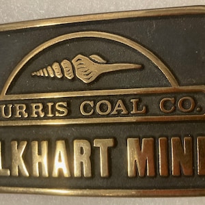 May include: A gold-colored metal belt buckle with a black background. The buckle features a stylized seashell design and the text "Turris Coal Co. Elkhart Mine".