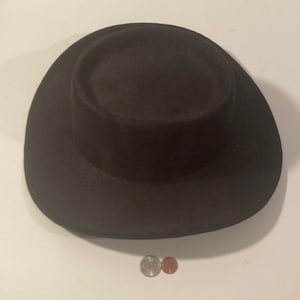 Pode incluir: Um chapéu fedora de feltro preto com uma aba larga. O chapéu está sobre uma superfície branca.