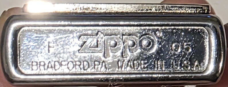 May include: Close-up of the bottom of a silver Zippo lighter. The text "Zippo" is prominently displayed, along with "BRADFORD PA. MADE IN U.S.A." and the number "05". The lighter has a shiny, reflective surface.