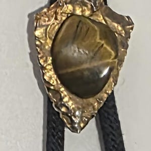 May include: A bolo tie featuring a gold-toned arrowhead pendant with a polished, oval-shaped stone. The pendant is attached to a black braided cord. The stone has brown and gold tones.