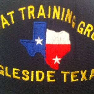 May include: A black baseball cap with yellow embroidered text that reads "Afloat Training Group Ingleside Texas". The text is in a curved arc around a blue and red embroidered image of the state of Texas.