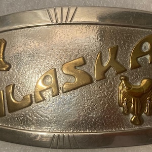 May include: Silver belt buckle with a gold inscription of "ALASKA" and two gold images, one of a cowboy boot and one of a saddle.