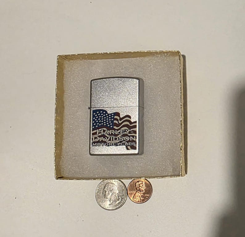May include: A silver Harley-Davidson lighter with a red, white, and blue American flag design. The lighter is in a small, square box with a quarter and a penny next to it.