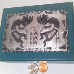 May include: A teal blue wooden box with a silver metal inlay depicting two Native American dancers. The dancers are in a stylized design with geometric patterns. The box is closed and sitting on a white surface.