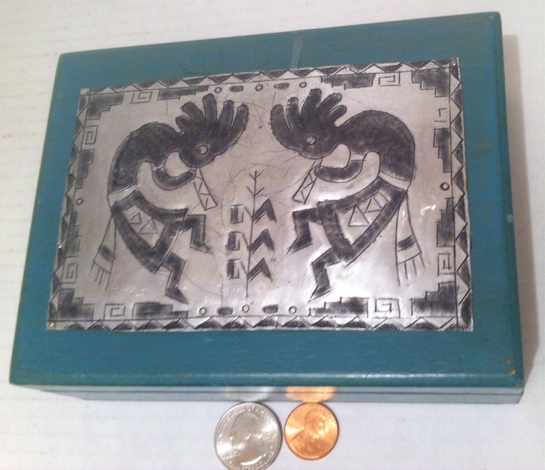 May include: A teal blue wooden box with a silver metal inlay depicting two Native American dancers. The dancers are in a stylized design with geometric patterns. The box is closed and sitting on a white surface.