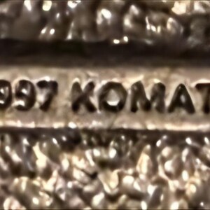 Op de afbeelding: Close-up van een metalen object met de tekst "&copy; 1997 KOMATSU" in reli&euml;f. Het object heeft een getextureerd oppervlak en lijkt een kleine, rechthoekige plaat of embleem te zijn.